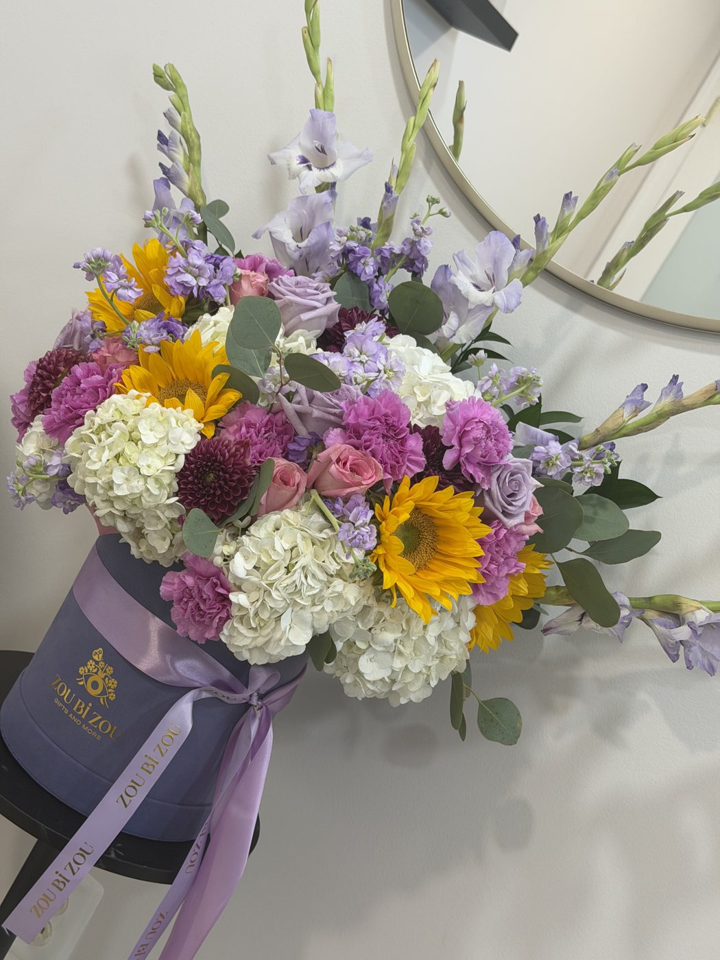 Lavender Zou Bi Zou hatbox of sunflowers, hydrangea, pink carnations, purple roses, stock and gladiolus