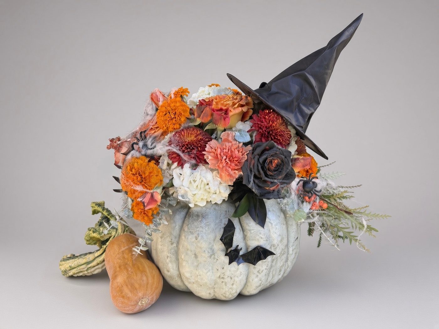 Halloween arrangement in a white pumpkin with marigolds, roses, black rose, spiderweb, bats and a witch hat