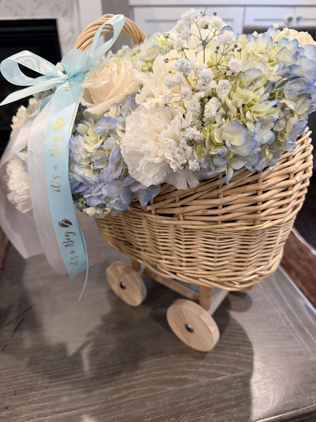 Wicker baby carriage filled with blue hydrangea, white carnations, roses and baby's breath with an It's a Boy ribbon