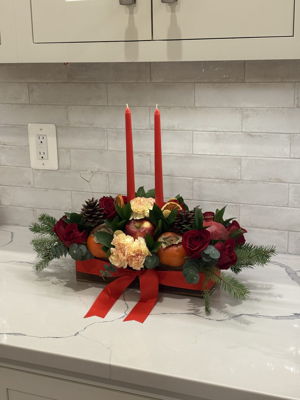 Holiday table centerpiece in a wooden box with red roses, persimmons, apple, carnations, pine and two red taper candles