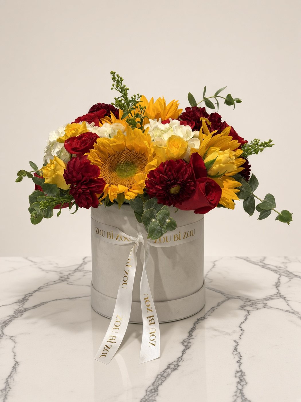 Grey velvet Zou Bi Zou hatbox of sunflowers, red roses, dahlias, yellow roses and hydrangea with a branded ribbon