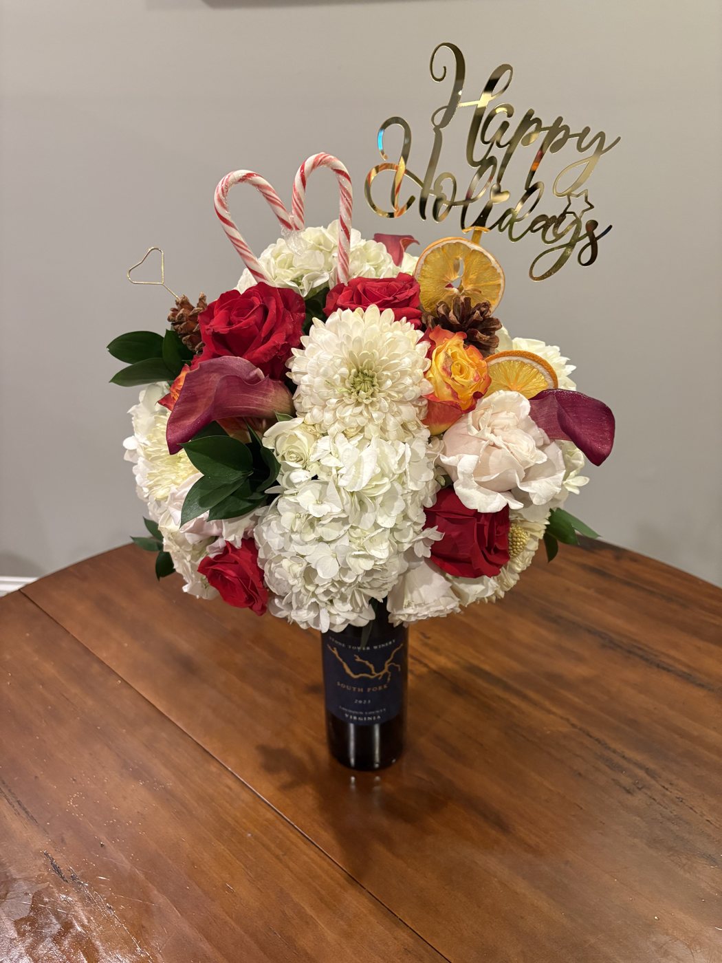 Holiday arrangement on a wine bottle with white hydrangea, red roses, calla lilies, candy canes, dried oranges and a Happy Holidays topper